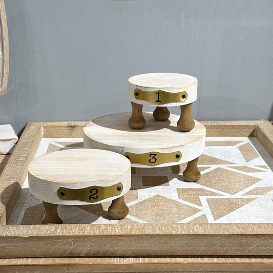Set of three white wooden display risers with numbered brass plates and sturdy legs, ideal for showcasing candles, plants, and decor items.