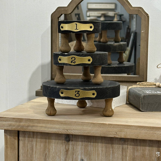 Set of three black round display risers with wooden legs, numbered 1 to 3 in gold plaques. Solid wood construction, perfect for showcasing decor items.