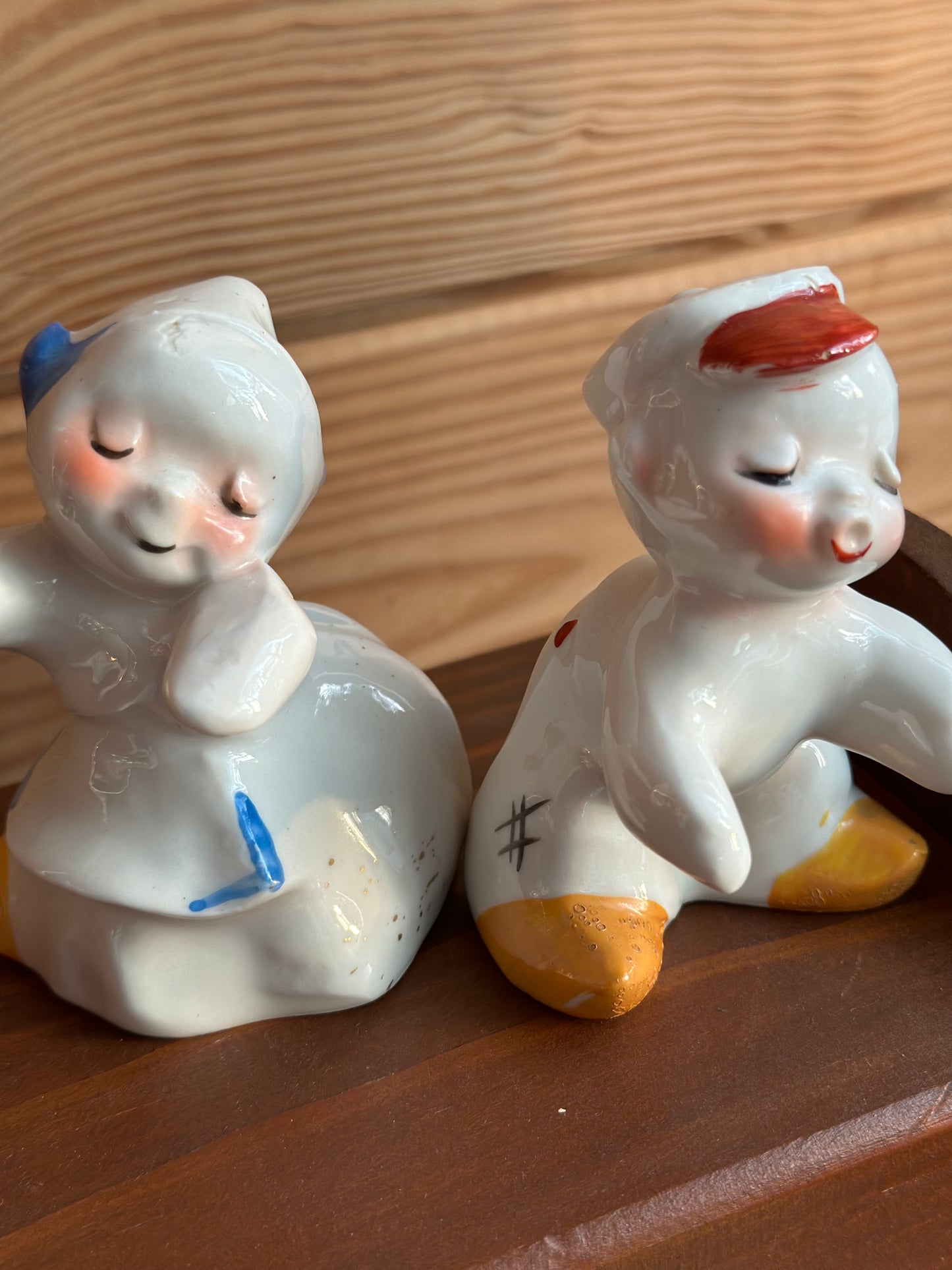 Ceramic baby boy and girl salt and pepper shakers with hand-painted details, sitting separately on a wooden surface. A charming vintage kitchen decor piece.