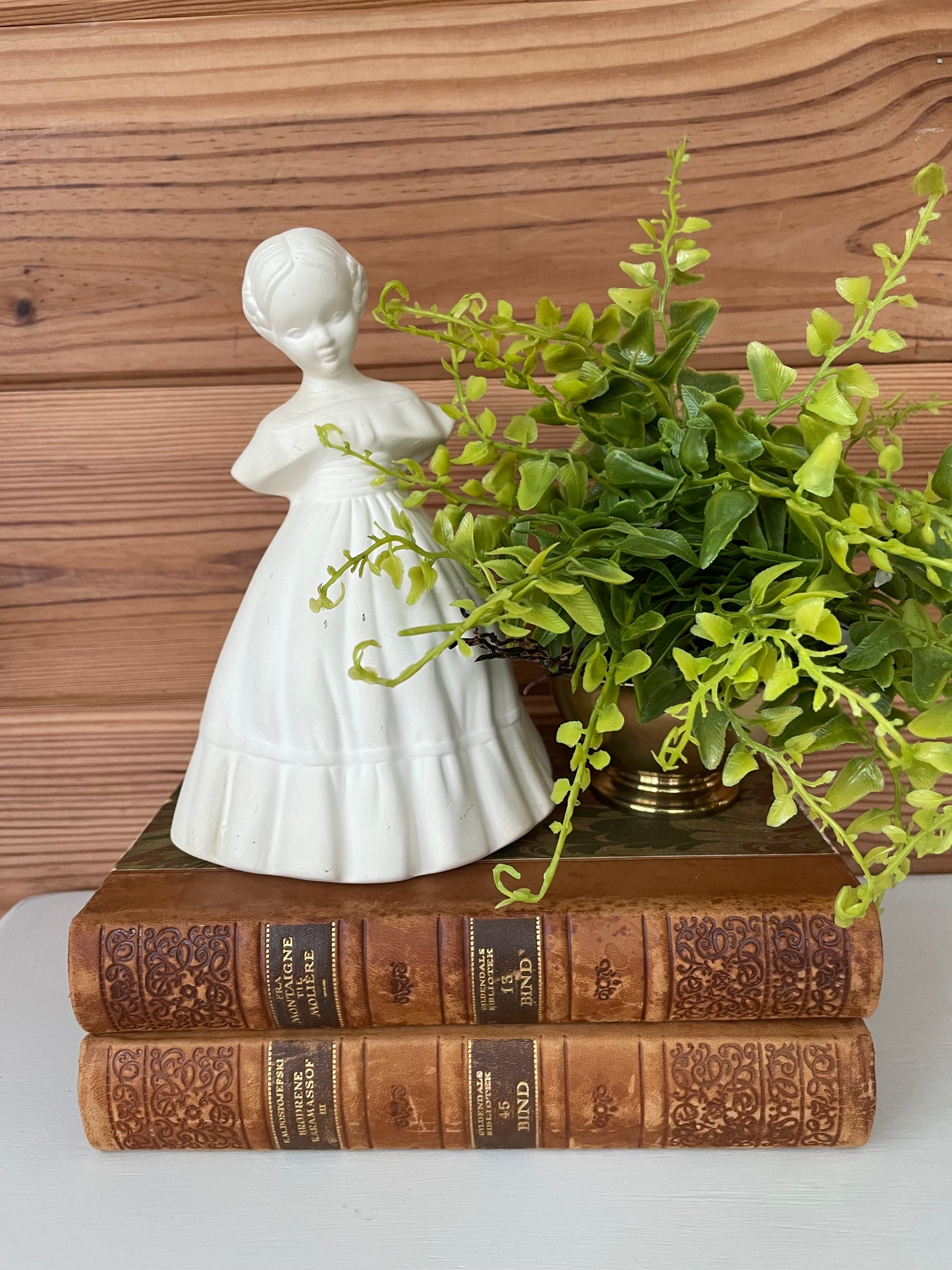 Elegant vintage ceramic girl figurine paired with antique books and a potted green plant. A charming decor accent for classic and modern interiors.