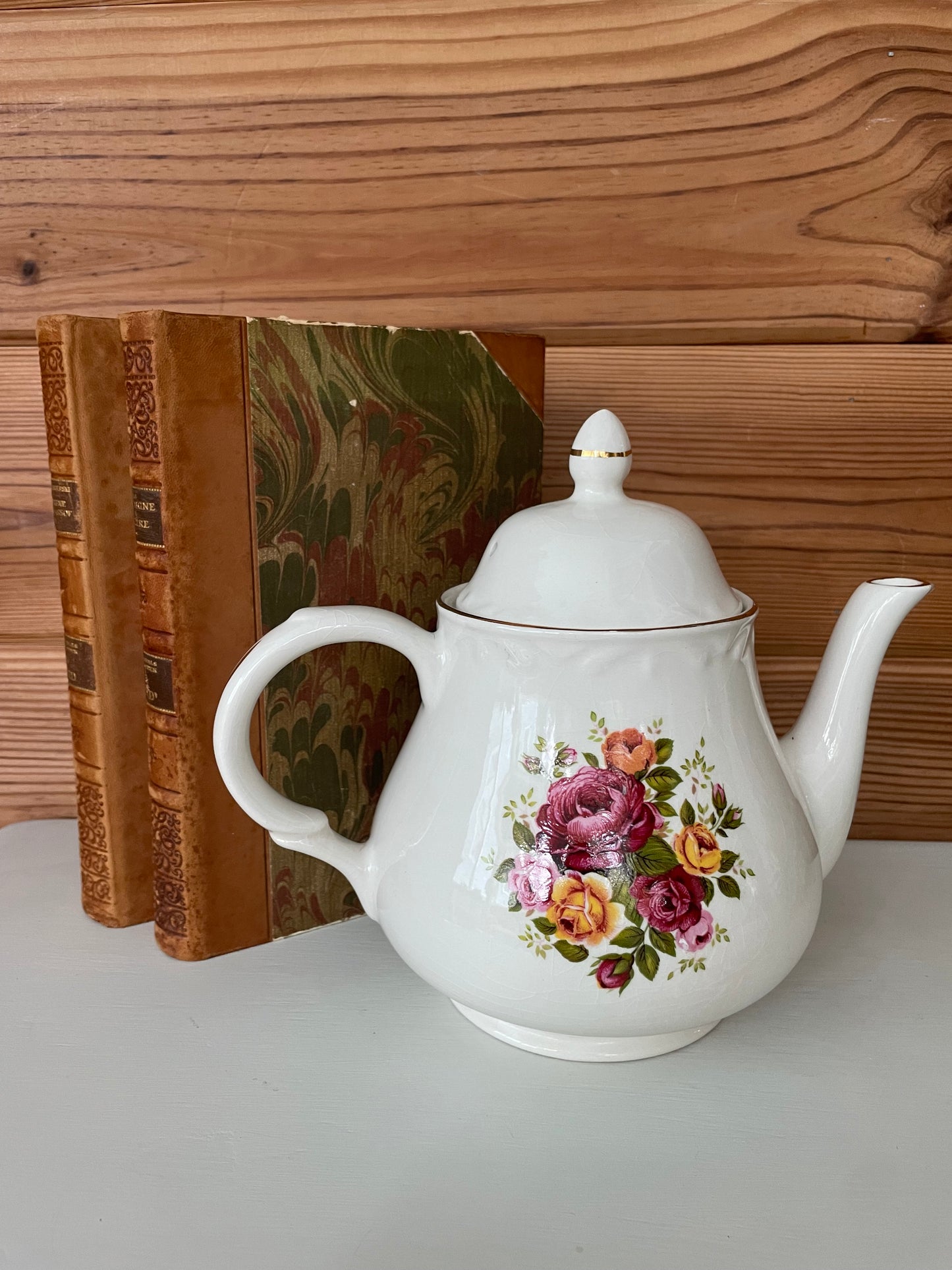 Charming vintage white teapot adorned with a floral rose pattern, set against antique books. A timeless piece for kitchen decor or tea gatherings. #6443
