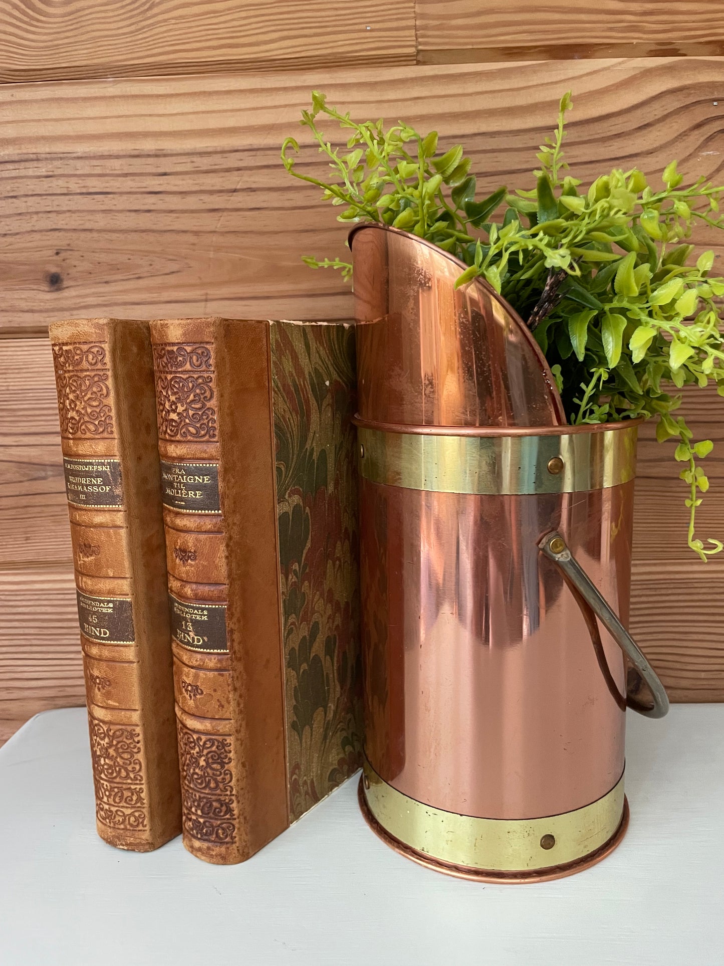 Vintage copper and brass fireplace match holder with a curved spout and handle, placed beside antique books and decorated with green foliage.