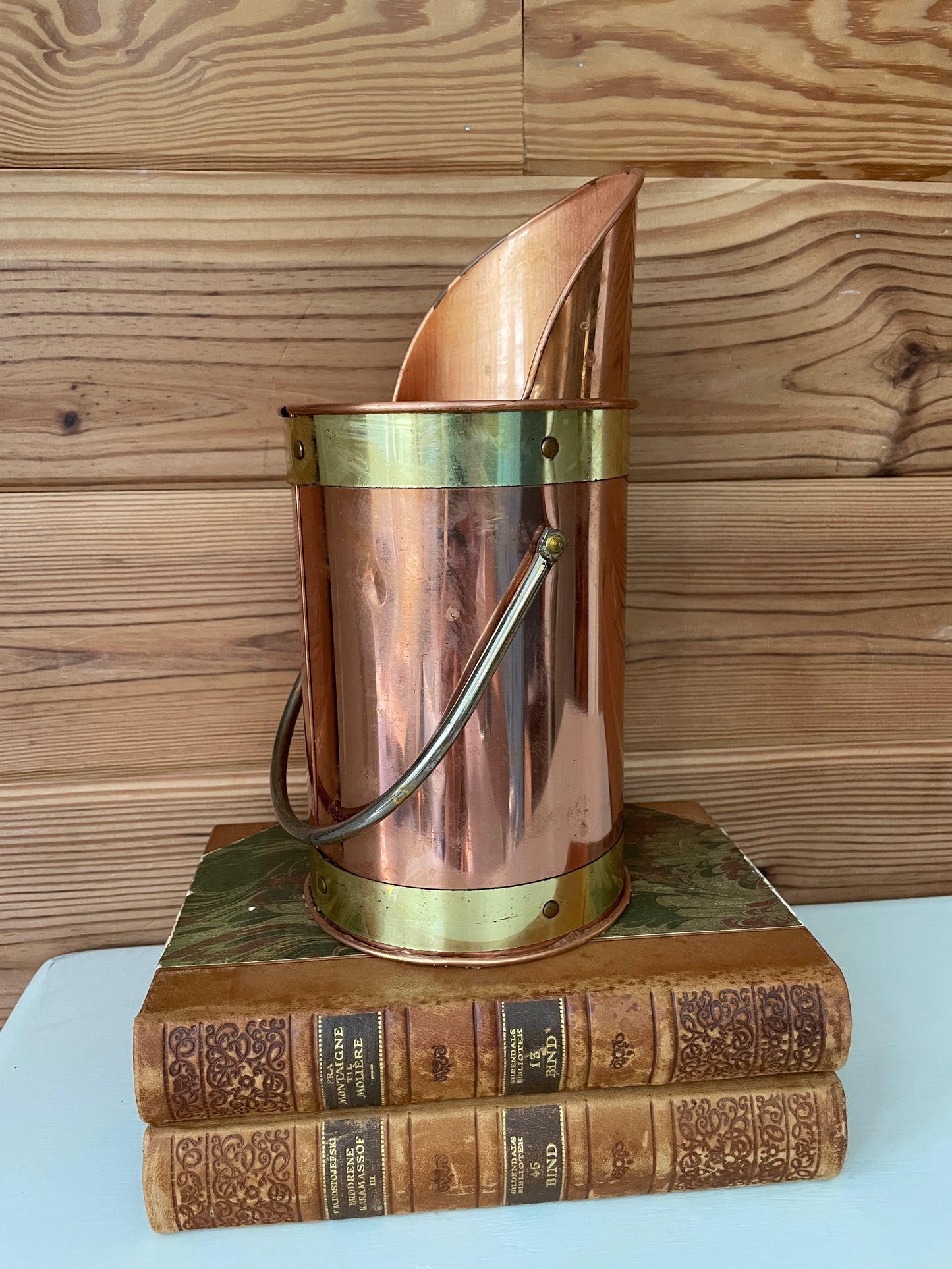 Stylish copper and brass fireplace matchstick holder with a curved spout and handle, displayed on antique books against a wooden backdrop.