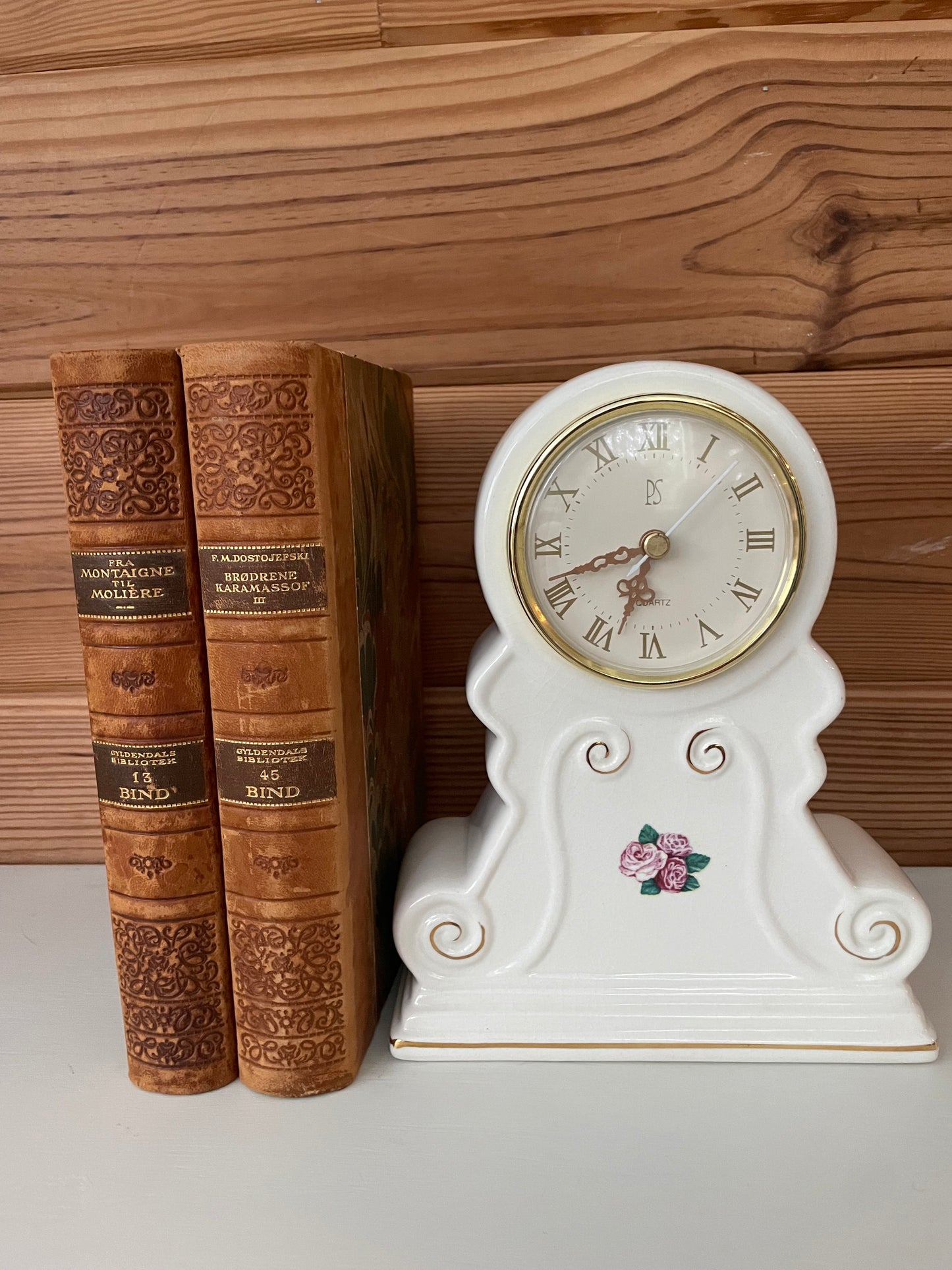 Vintage white porcelain mantel clock with gold-rimmed Roman numeral dial, adorned with delicate pink floral accents, placed beside antique books.