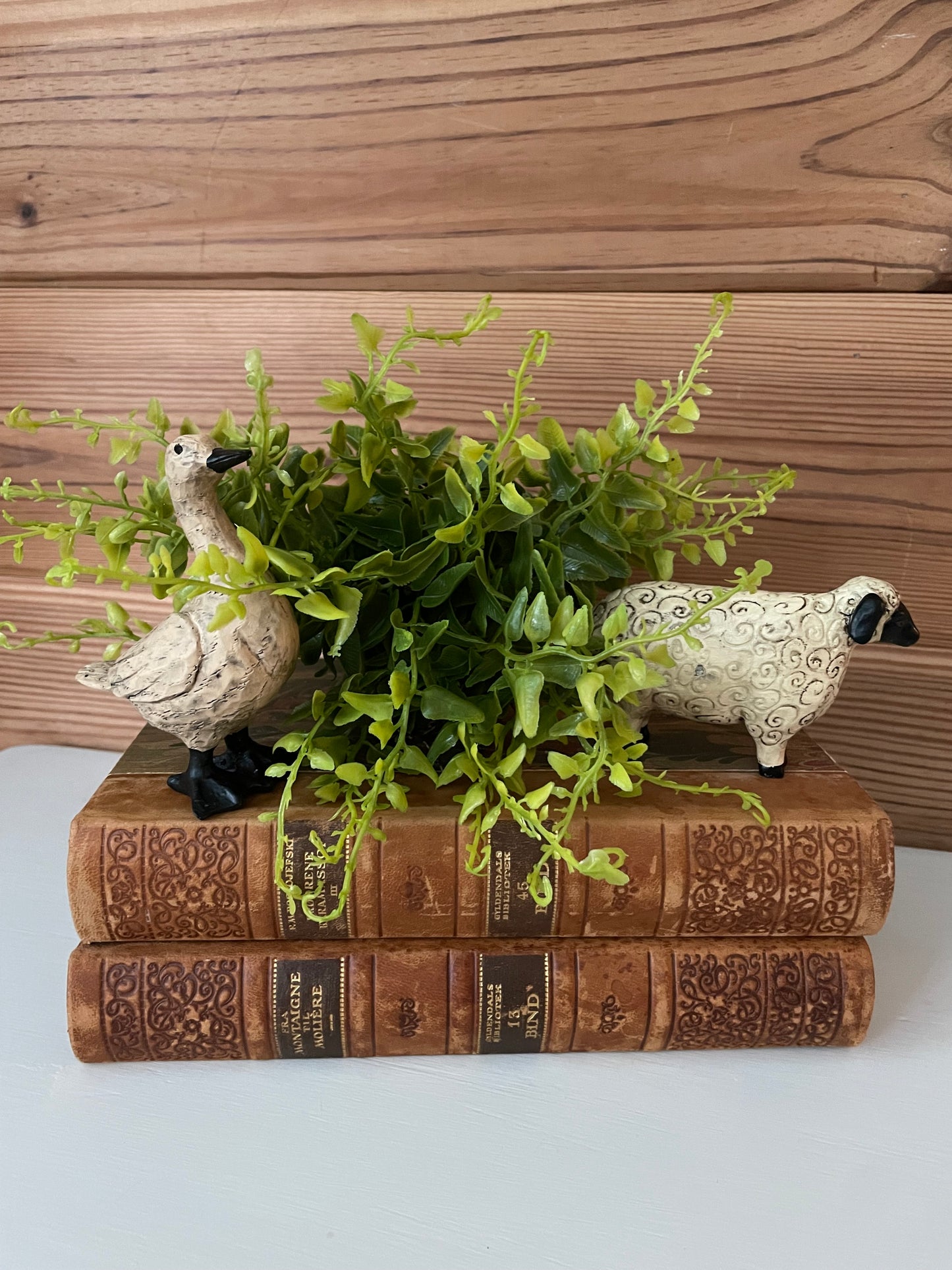 Goose and sheep resin figurines placed on antique books with greenery. Charming farmhouse-style decor for home and garden spaces.