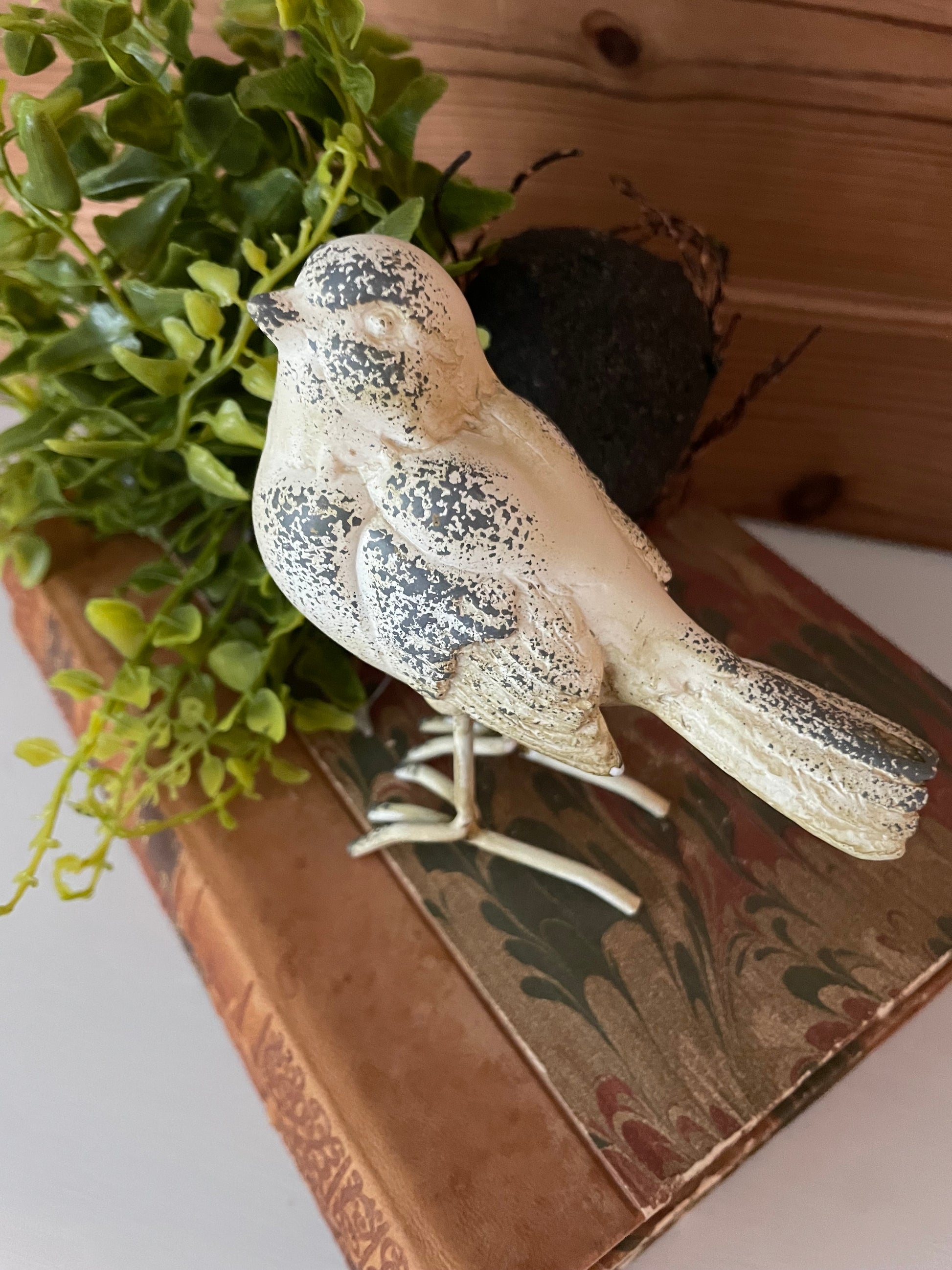 Close-up of white/grey distressed resin metal bird decor on antique books with greenery. A perfect decorative piece for home and garden styling.