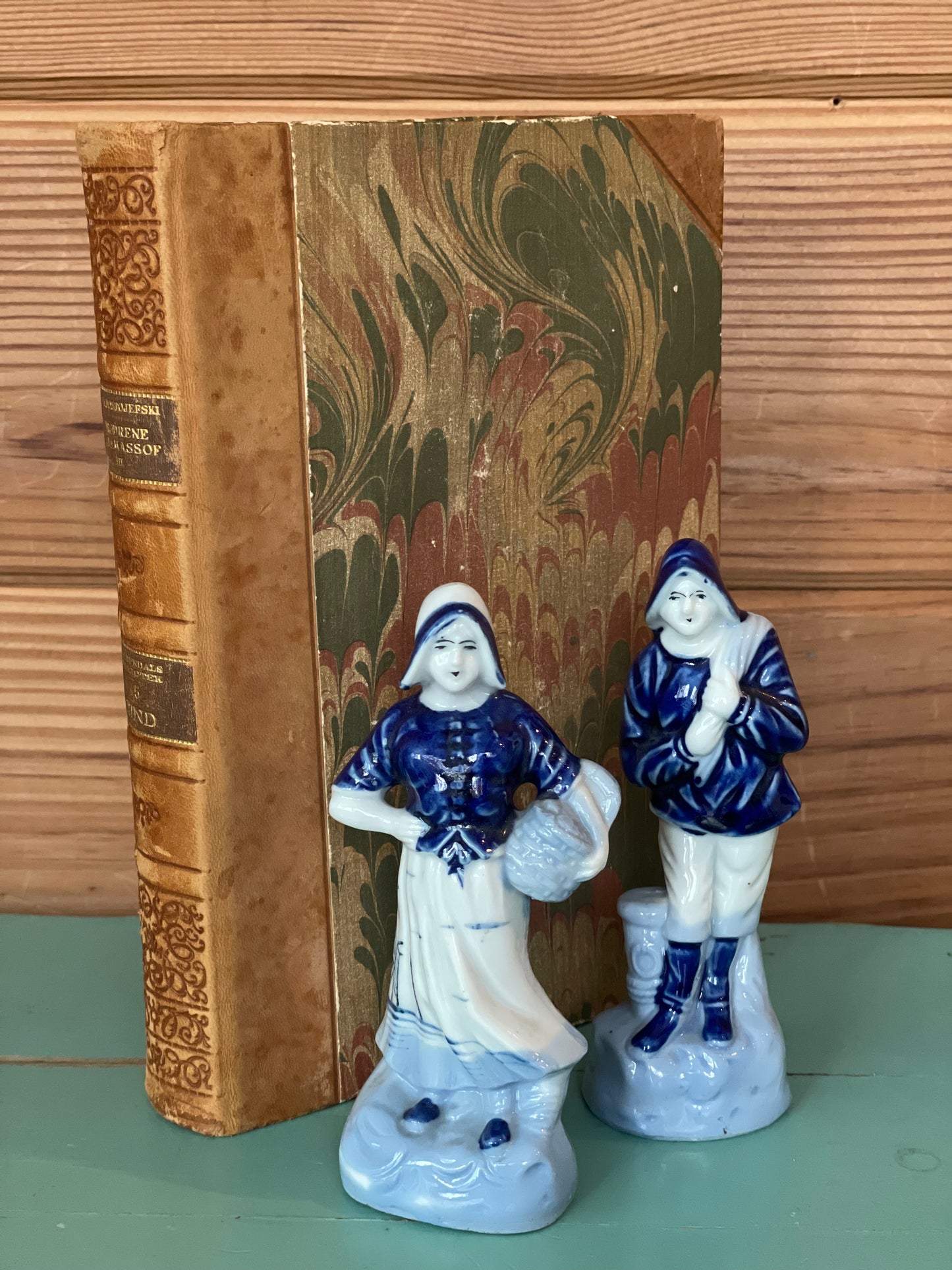 Vintage Blue Delft porcelain figurines of a Dutch woman with a basket and a fisherman, hand-painted in classic blue and white, displayed beside an antique book.
