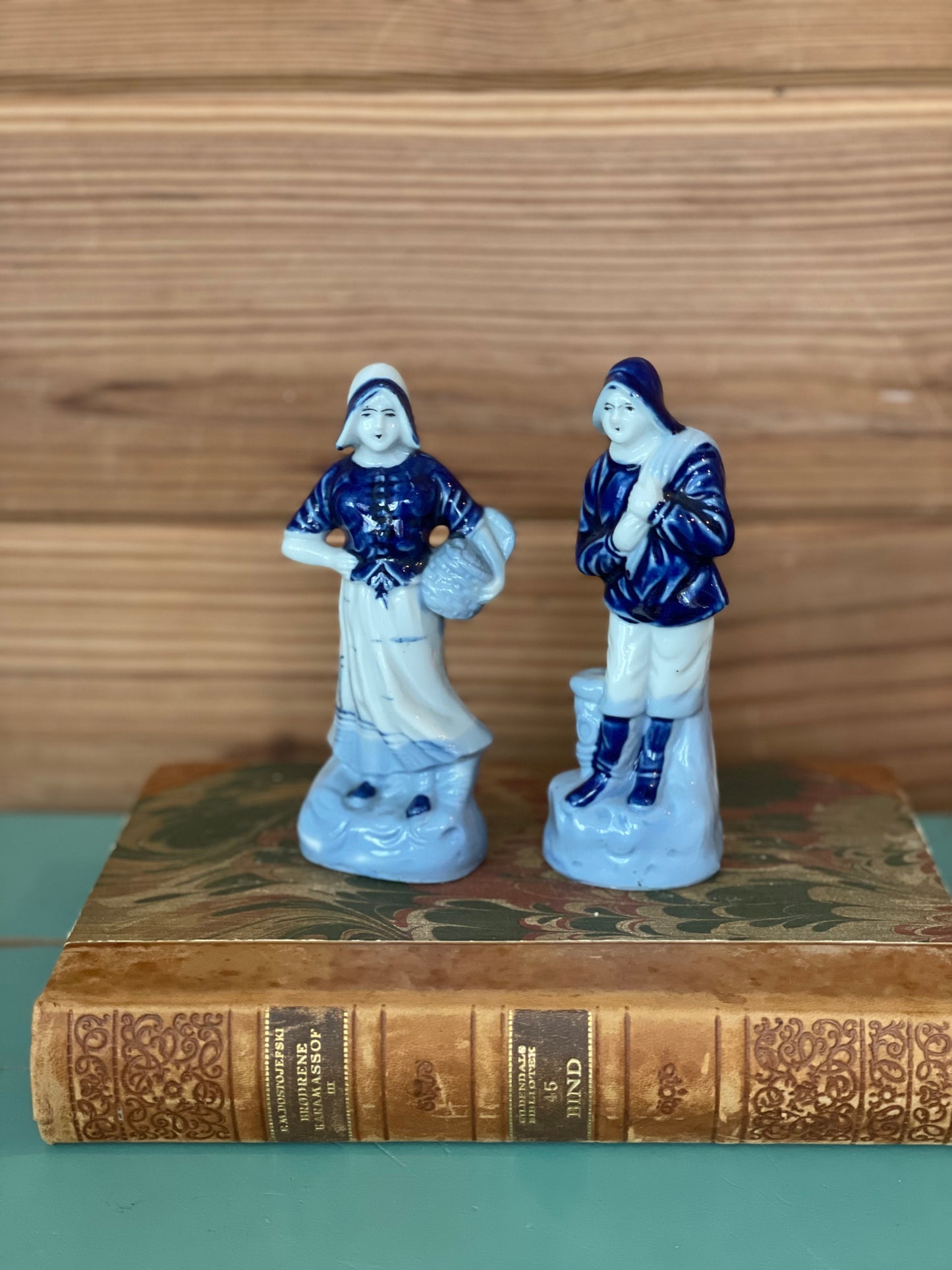 Pair of Blue Delft porcelain figurines featuring a Dutch woman and fisherman, set atop an antique book with a marbled cover, perfect for collectors.