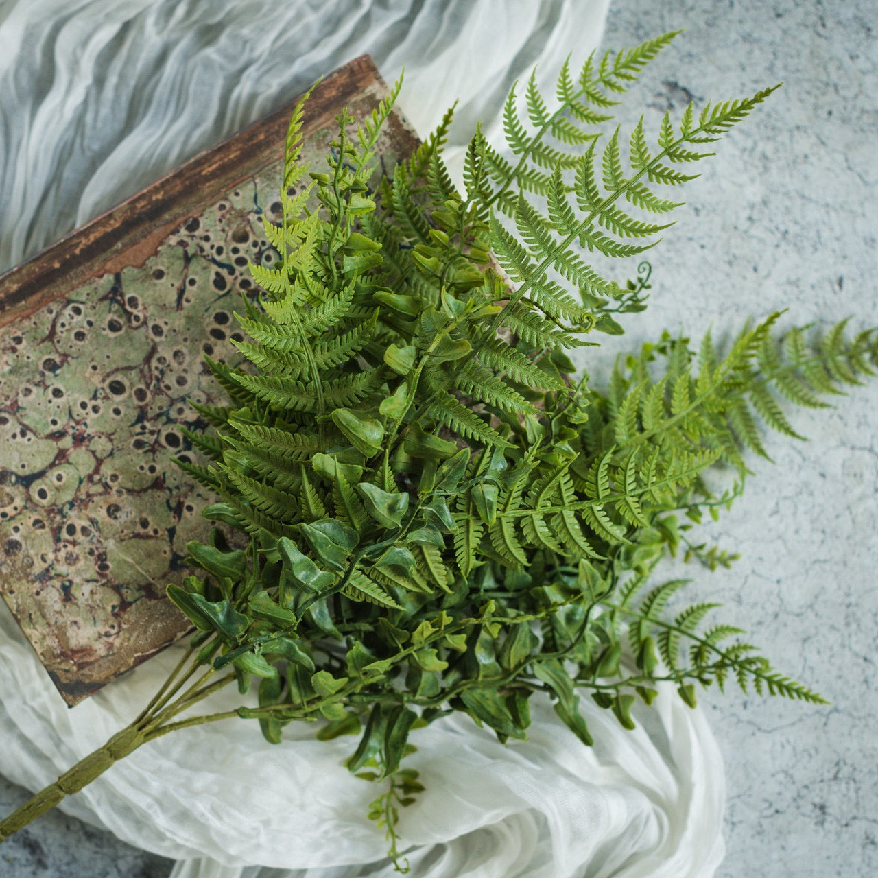 15-inch Buckler Fern Bush with realistic faux greenery, placed beside an antique book on a textured surface. Perfect for home and garden decor.