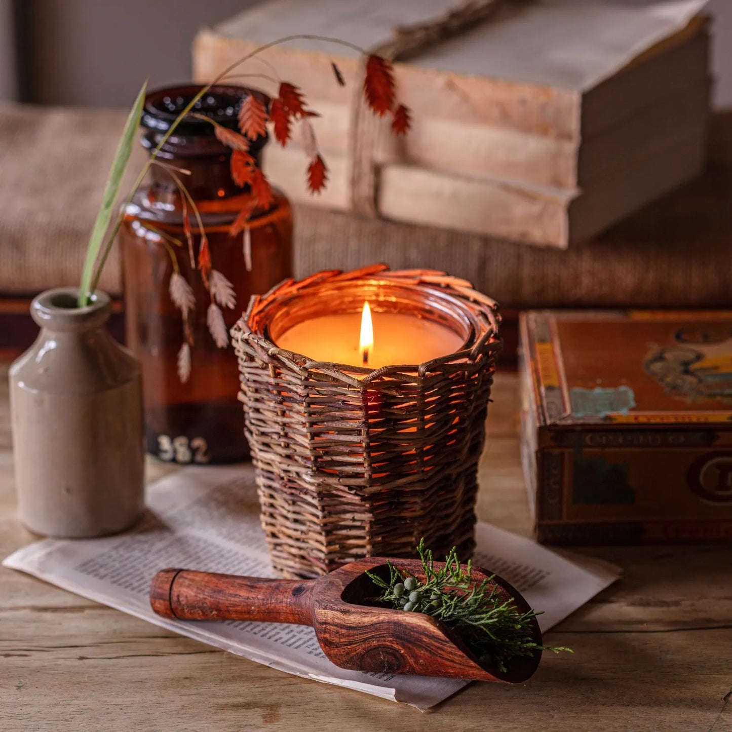 Porch View Home Hickory Tobacco & Tweed Candle in a woven willow holder, emitting a warm glow. Rustic decor with vintage books and amber glass bottle.