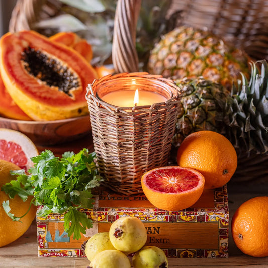 Porch View Home - Havana Candle in a woven holder, surrounded by fresh pineapple, papaya, oranges, and herbs. Features a lemon pineapple scent.