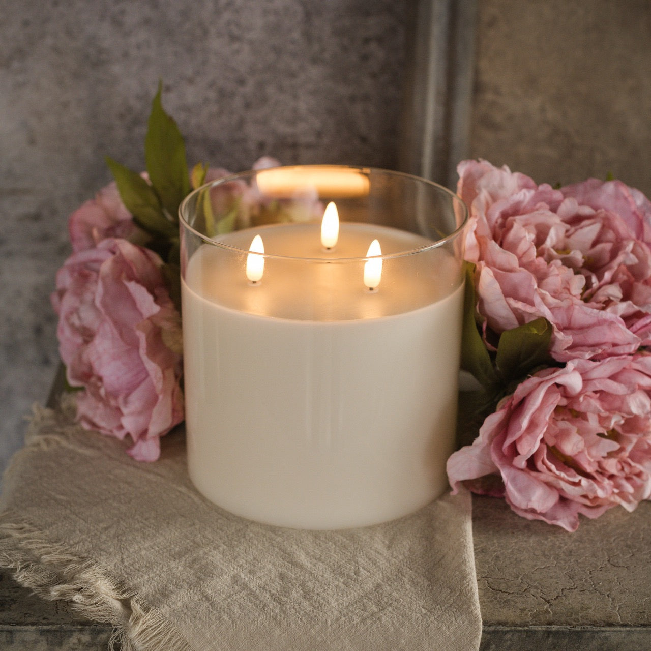Glass 3 Wick 3D Flame Candle - 6-inch with realistic flickering flames, set among pink peonies on a rustic surface. Elegant flameless home decor.