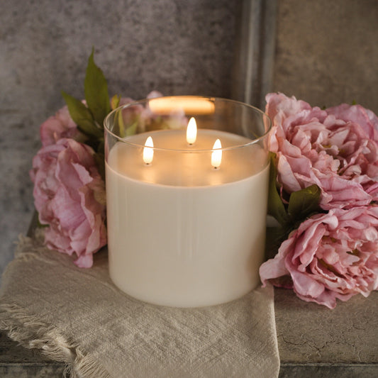 Glass 3 Wick 3D Flame Candle - 6-inch with realistic flickering flames, set among pink peonies on a rustic surface. Elegant flameless home decor.