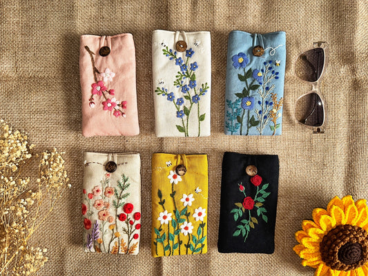 Set of six flower embroidered soft linen padded glasses cases in various colors with button closures, displayed on burlap fabric with sunglasses.