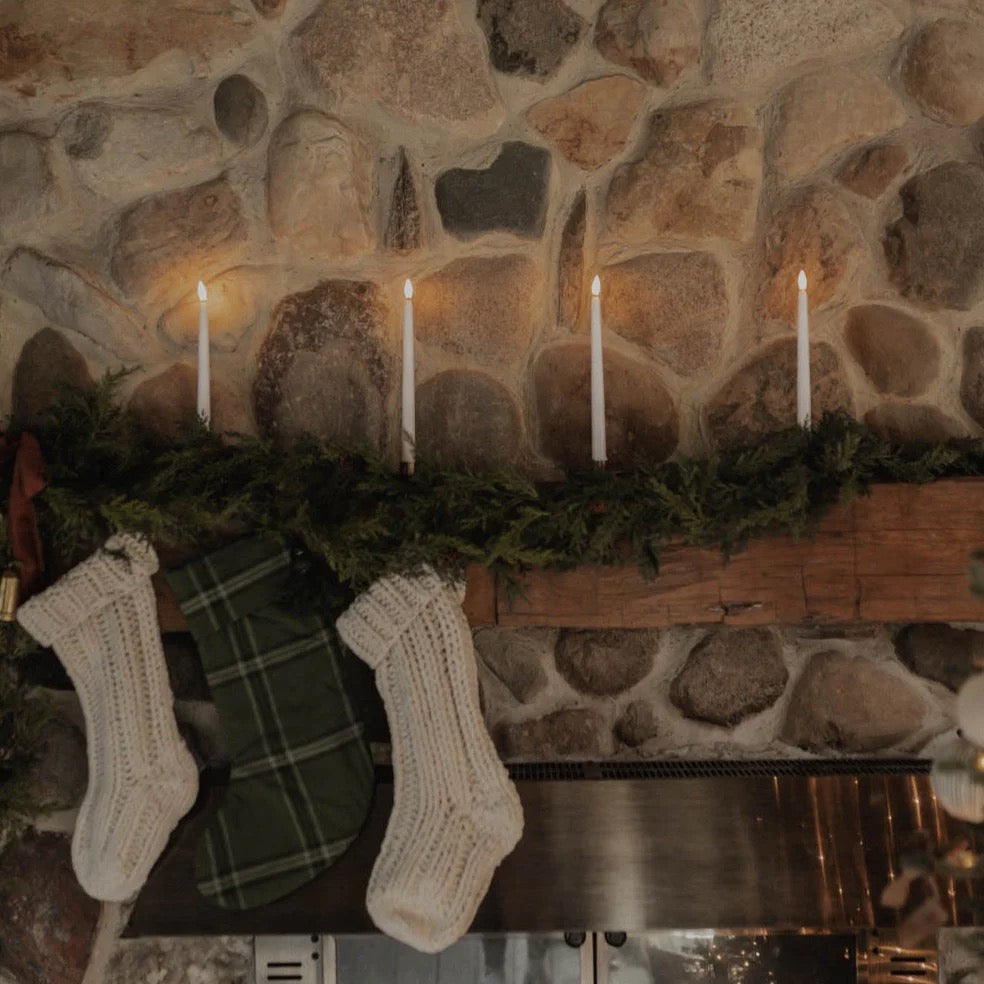 Antique Candle Co. flameless taper candles elegantly placed on a rustic fireplace mantel with holiday stockings and greenery.