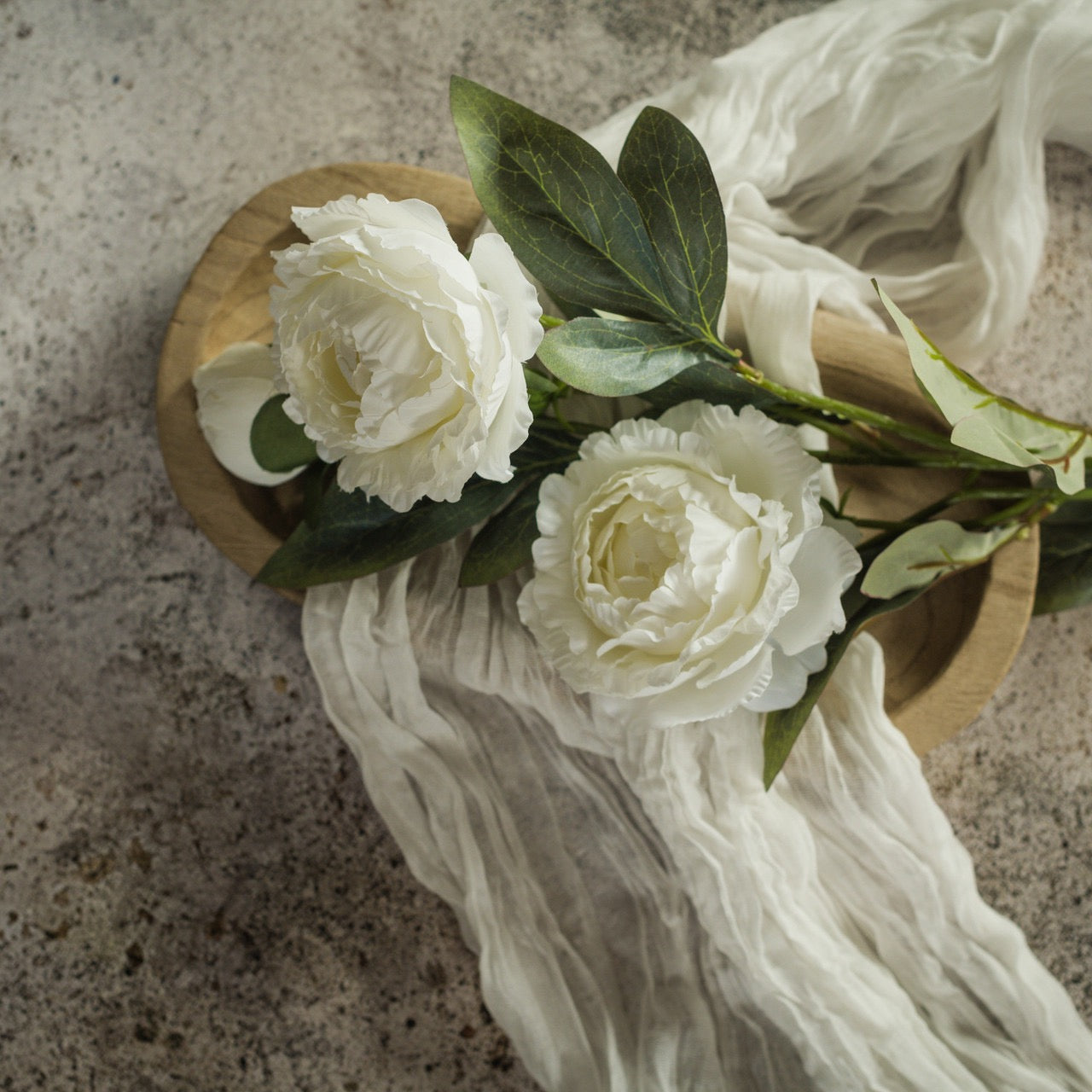 Elegant 27.5-inch white peony spray with lifelike ruffled petals and lush green leaves, artfully arranged on a wooden tray with soft fabric draping.
