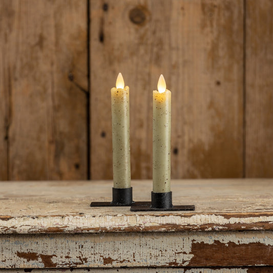 Set of two 6.75-inch moving flame green taper candles with a rustic finish, placed on black metal holders against a wooden backdrop.