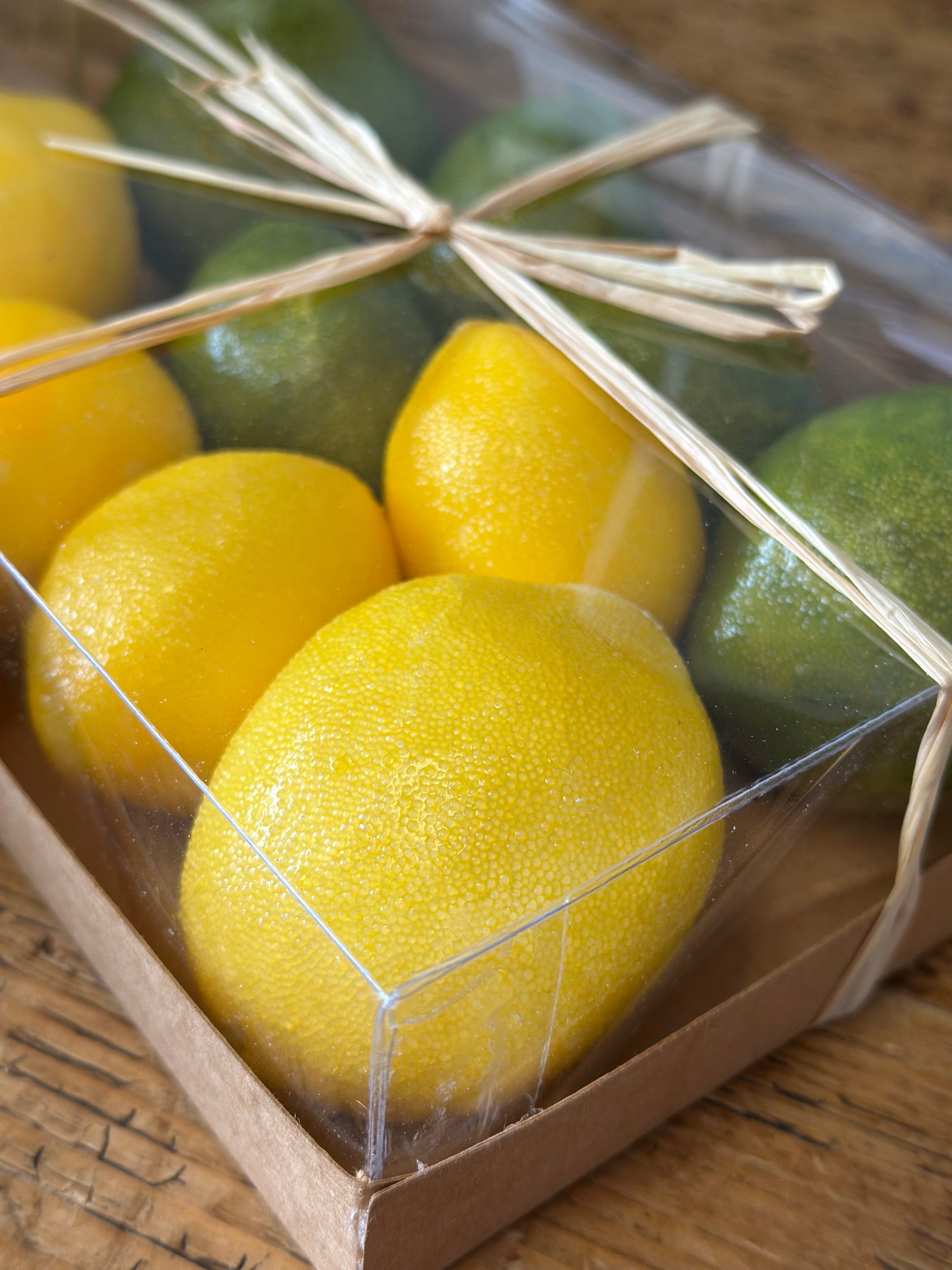 Close-up of faux lemon and lime set in a transparent box tied with raffia ribbon. Realistic artificial citrus fruits ideal for decorative displays and crafts.