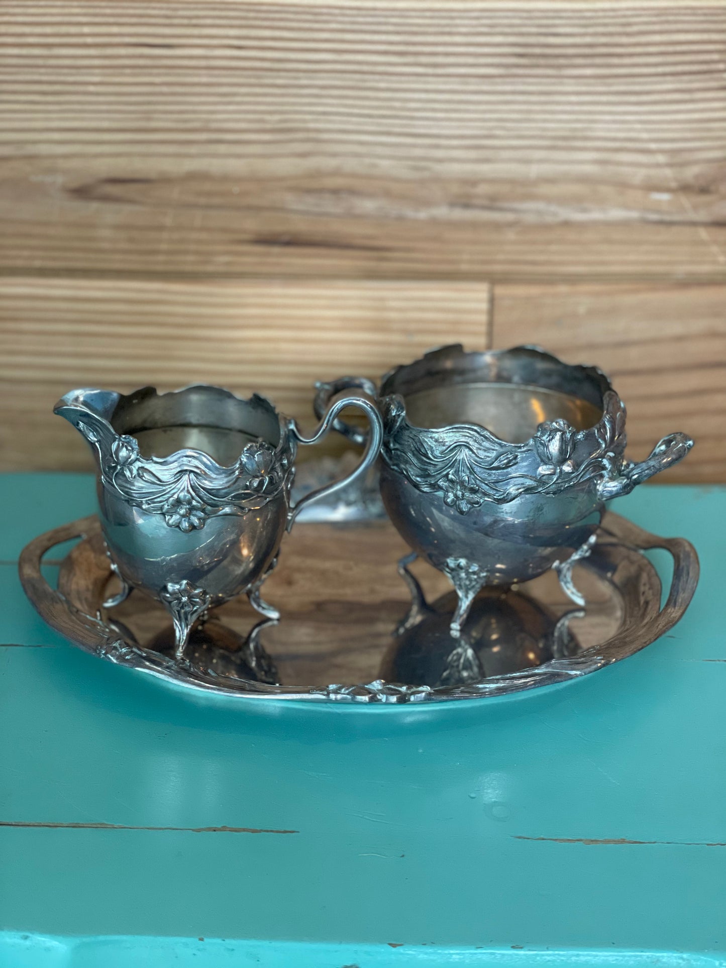 Vintage silver-plated creamer set with ornate floral detailing, including a tray, creamer, and sugar bowl. A timeless addition to any elegant table setting.