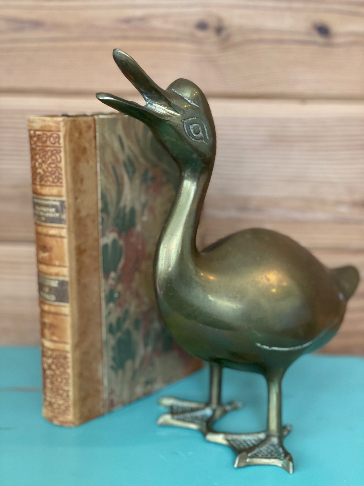 Vintage brass duck decor with an open beak, standing beside an antique book. A timeless home accent adding classic elegance to any space.