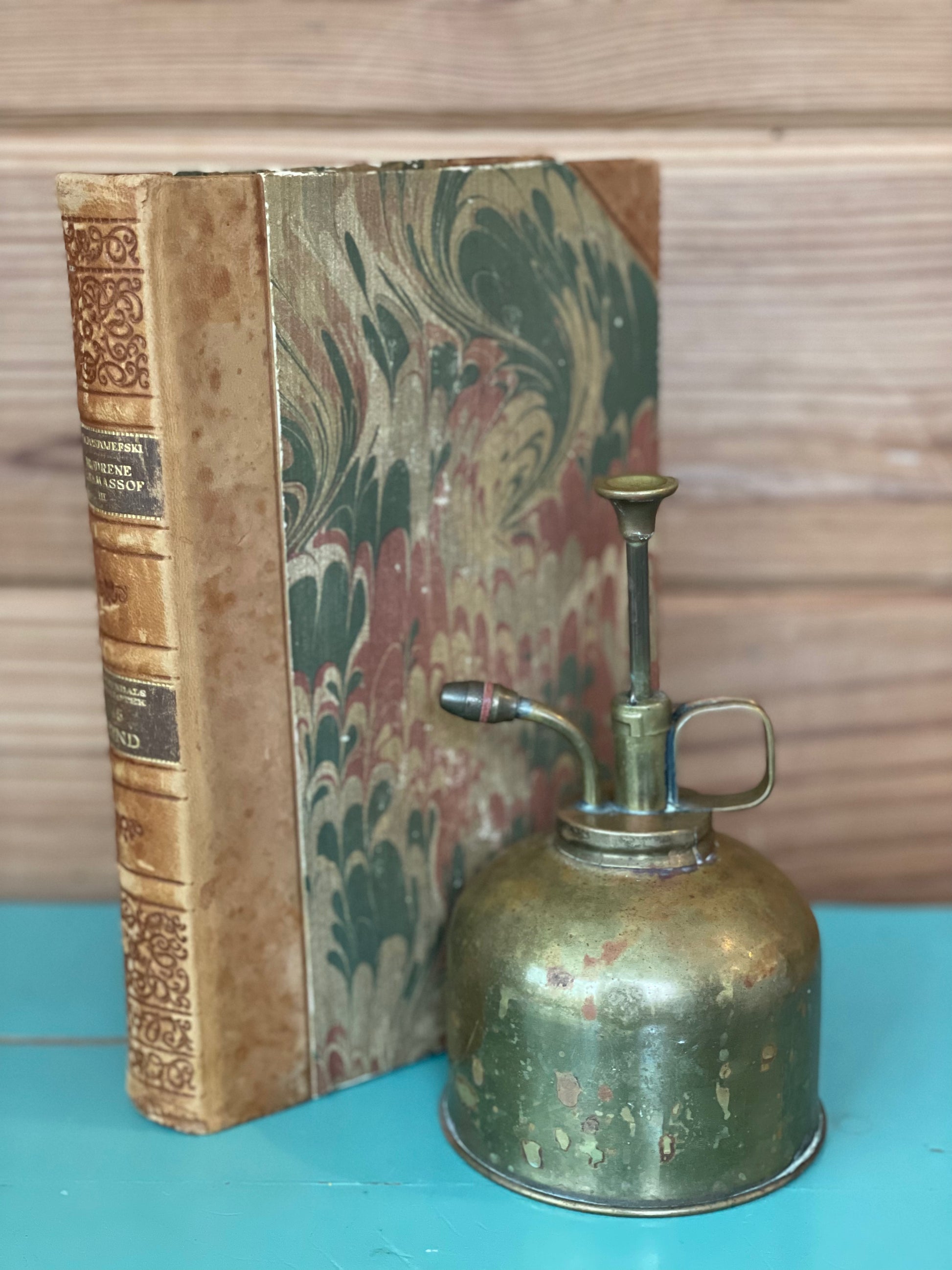 Antique brass plant mister with a patina finish, leaning against a vintage book. A charming addition to home decor and garden accessories.