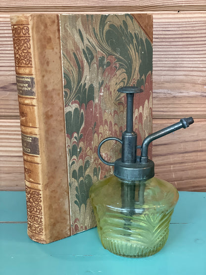 Vintage green glass mister bottle with a black metal pump, placed beside an antique book. Ideal for plant care and home décor with a retro charm.