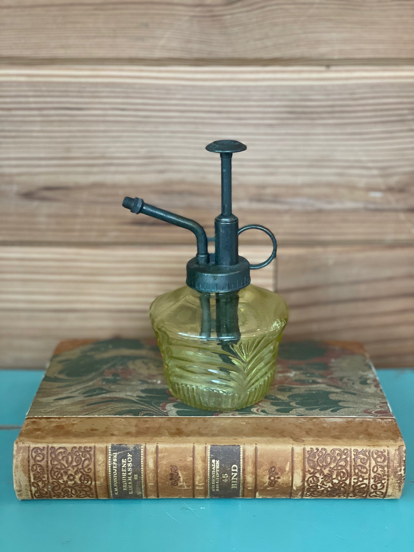 Vintage green glass mister bottle with a black metal pump, elegantly displayed on an antique book. A stylish and functional piece for plant care and décor.