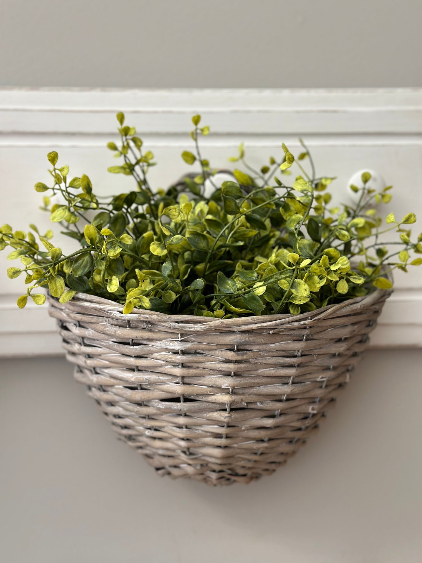 Gray wicker storage basket hanging on a white hook, filled with green artificial plants. A stylish and functional home decor piece.