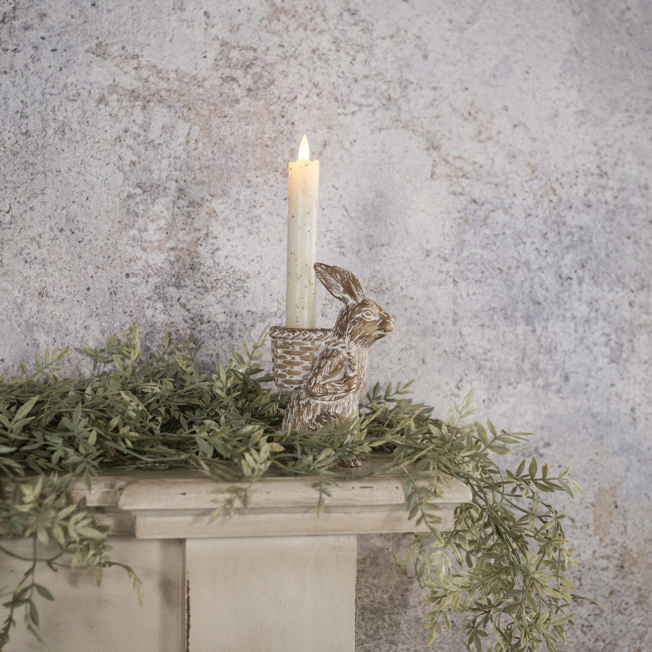 Charming bunny taper candle holder with a lit candle, displayed on a mantel with greenery. Ideal for Easter and cozy home decor.