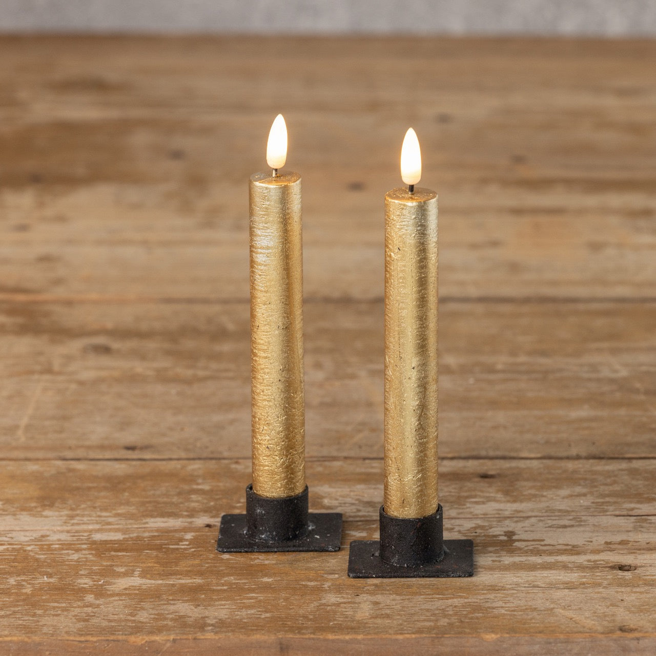 Pair of 3D flame gold taper candles on black metal holders, set on a wooden table. Flameless design offers a realistic glow for home decor.