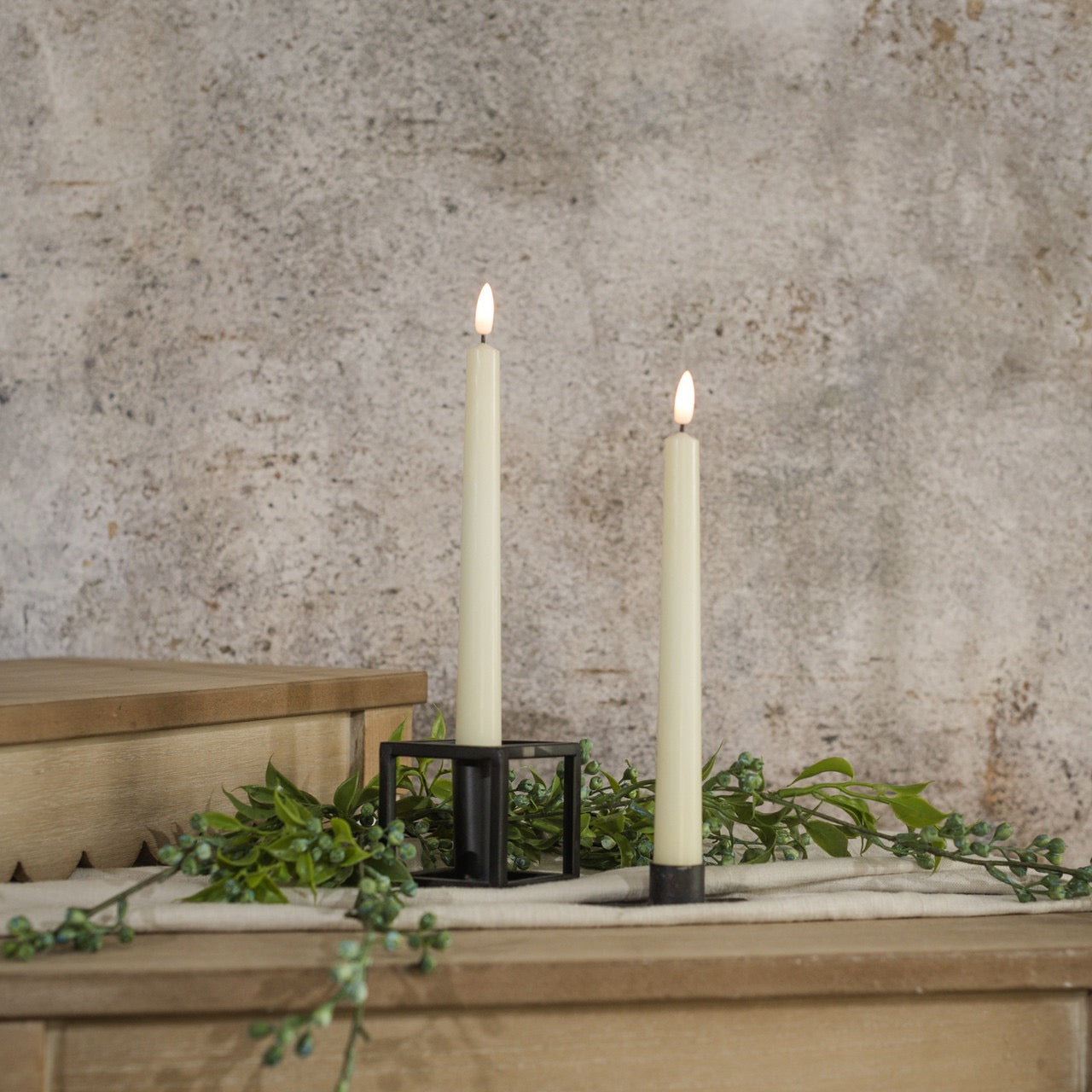 Set of 2 elegant 9.5-inch cream taper candles with 3D flame effect, placed in black holders on a wooden surface with greenery for a cozy ambiance.