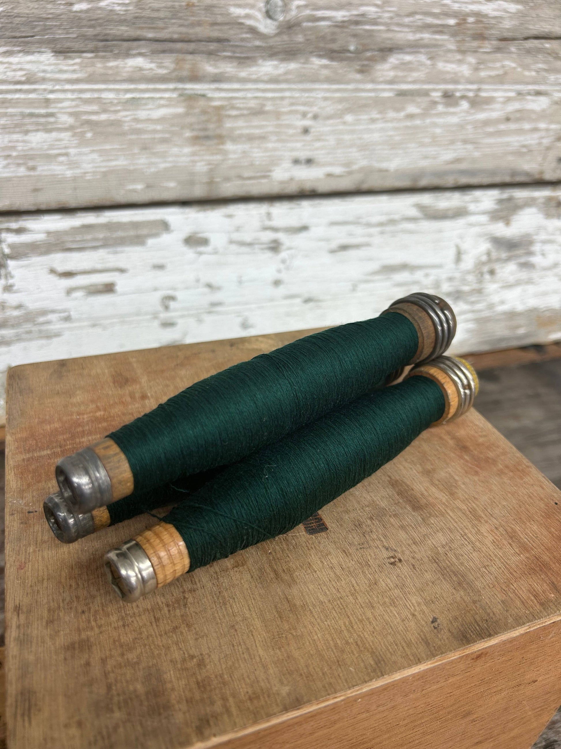 Dark green yarn quills with metal tips, wrapped in fine thread, resting on a wooden surface. Classic home decor piece for vintage-inspired interiors.